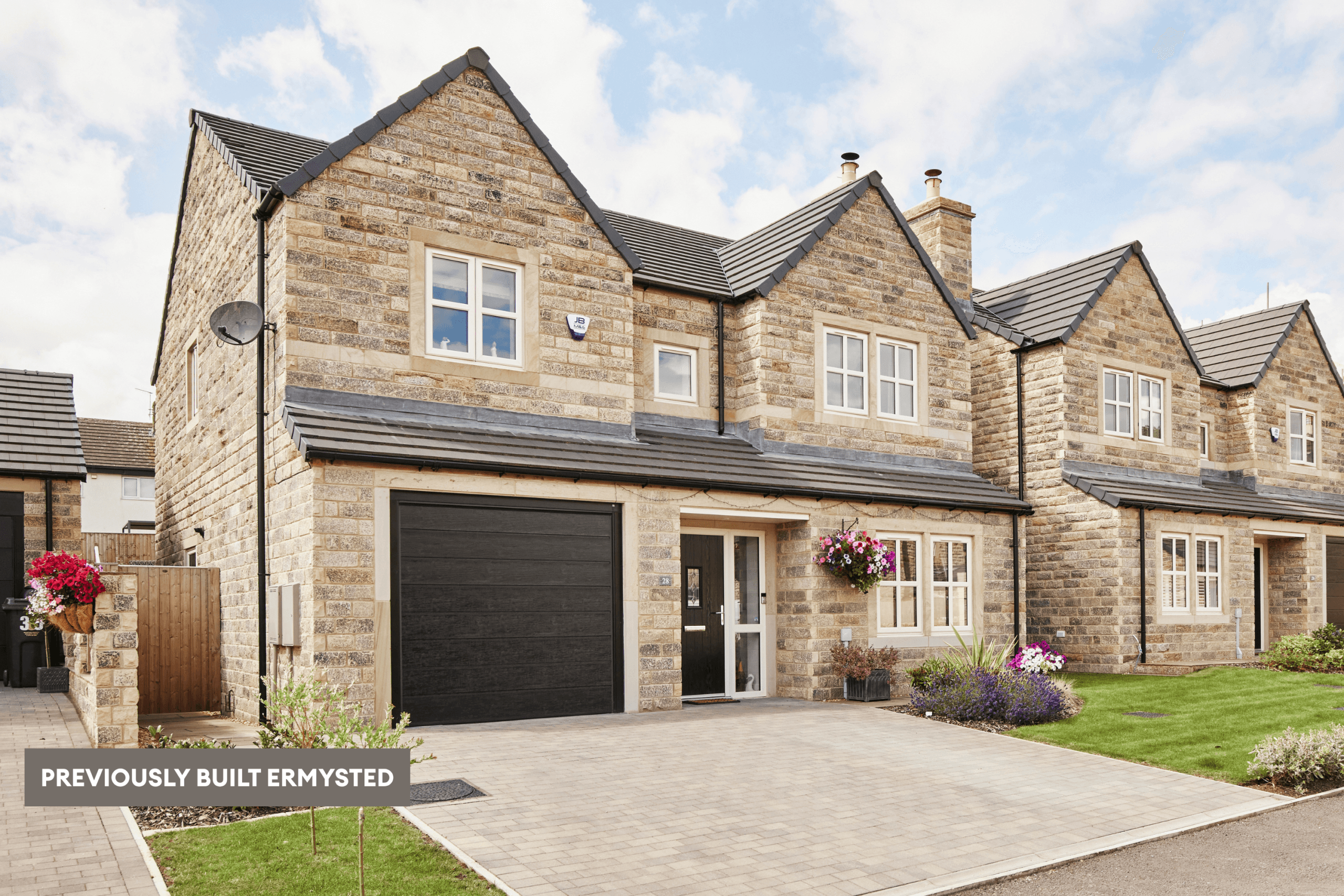 Previously built Ermysted home viewed from an angle, showing the natural stone exterior, integral garage, paved driveway and neighbouring homes.