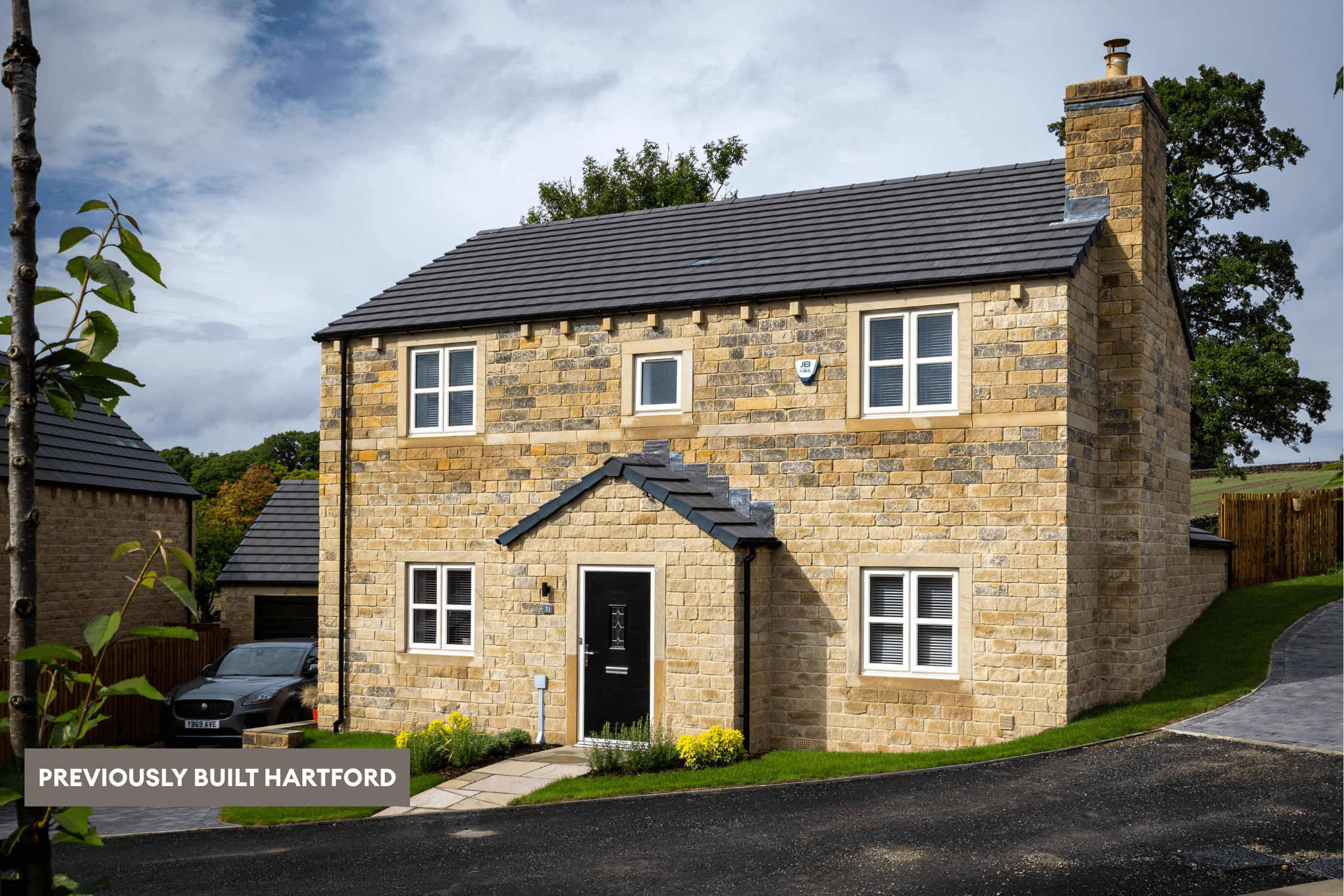 Previously built Hartford detached home with natural stone exterior, black front door, chimney, white-framed windows and sloping driveway.