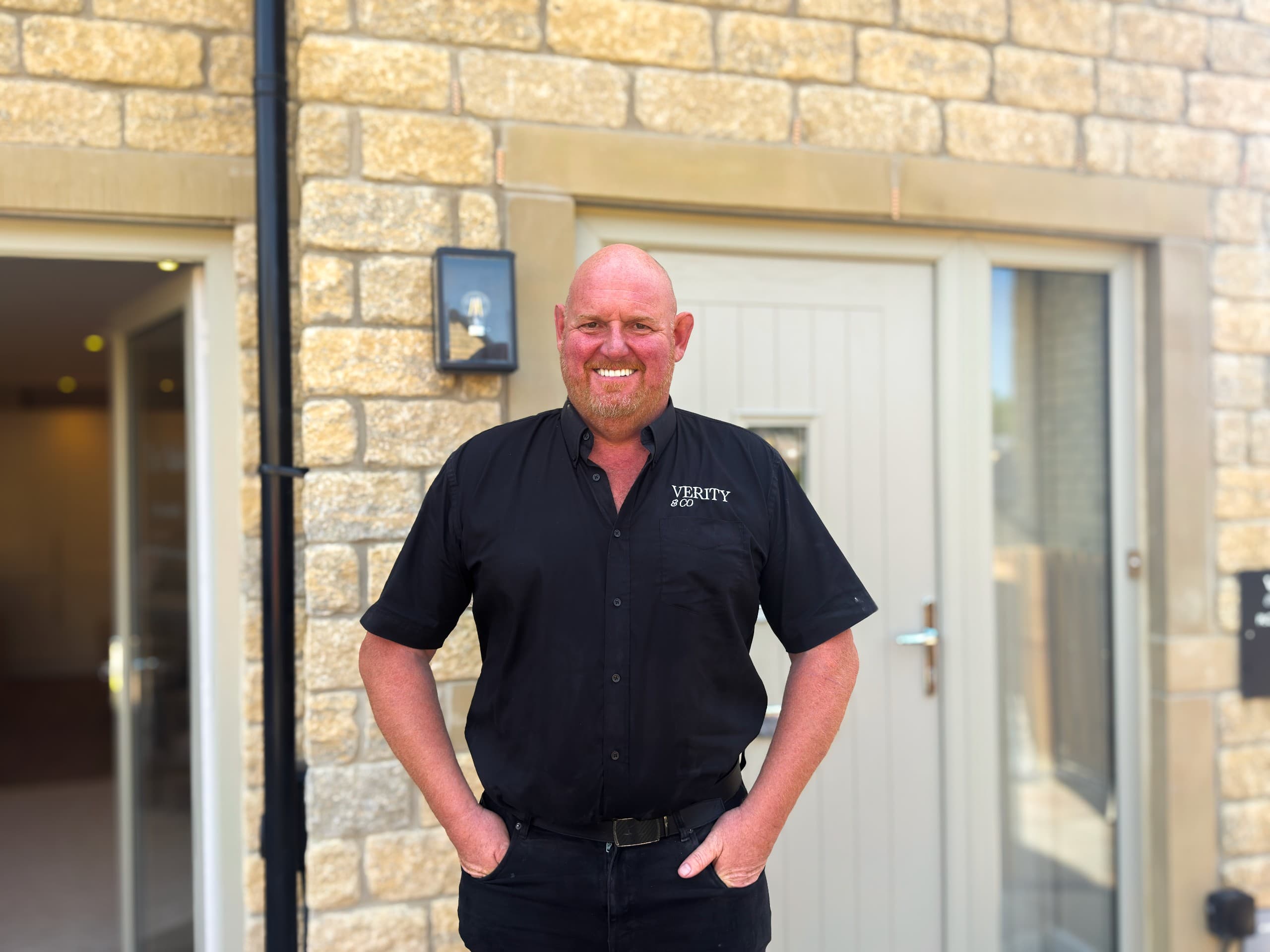 A smiling Verity & Co team member wearing a black branded shirt, standing outside a stone-built home with his hands in his pockets.
