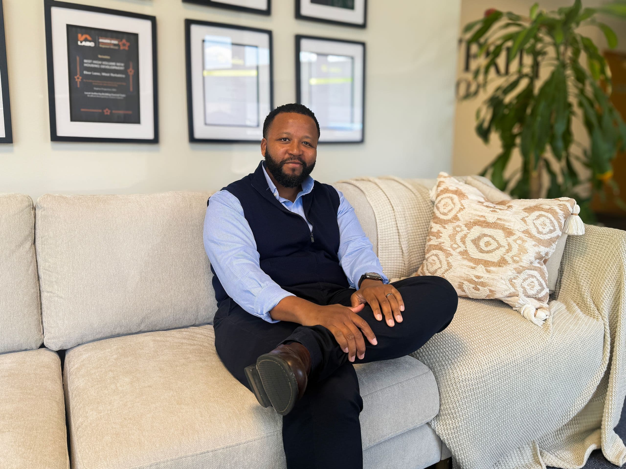 Maison Mokoena seated on a neutral sofa with his legs crossed, looking at the camera, with framed industry awards and certificates displayed behind him.