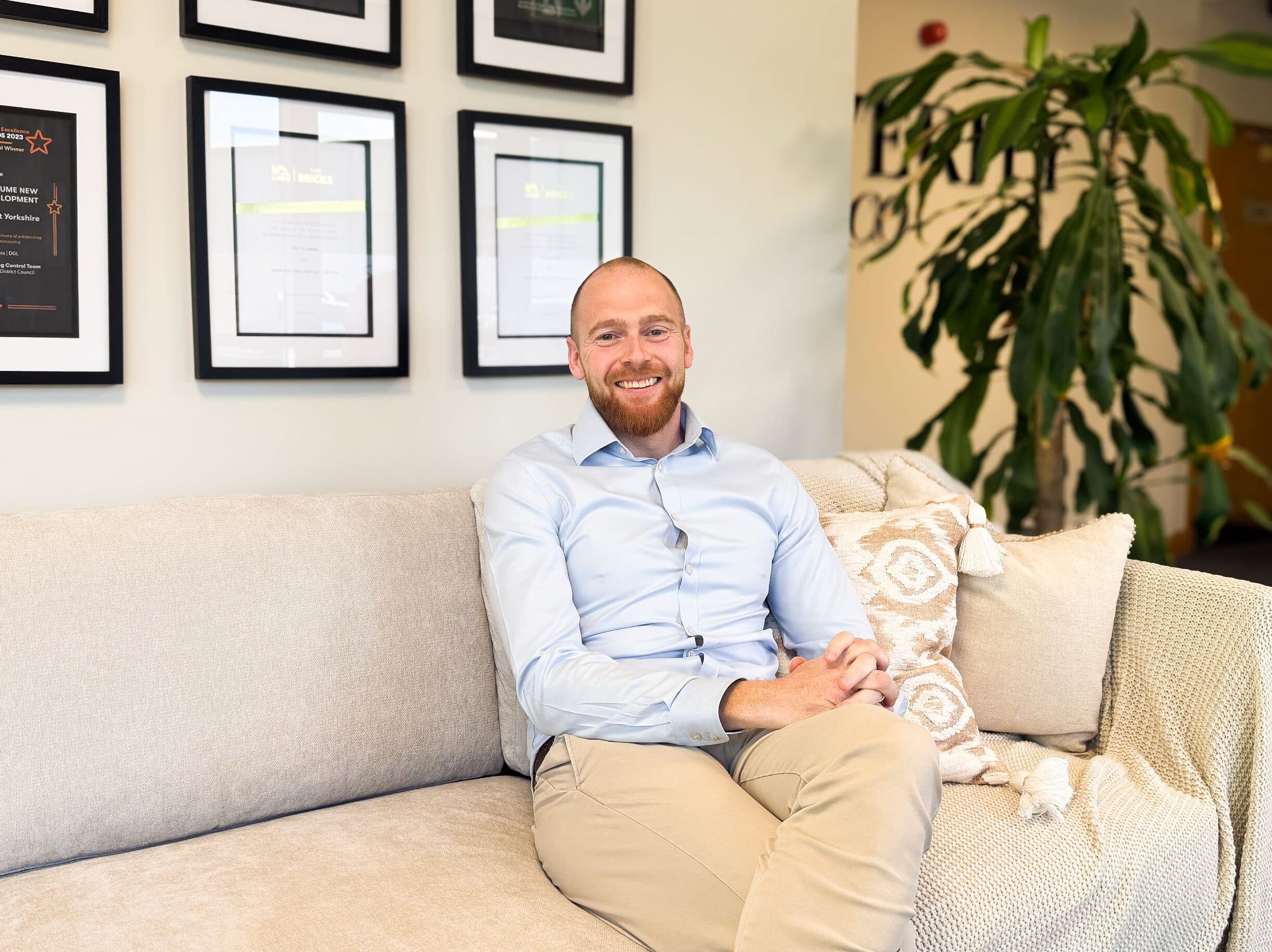Tom Pardey seated on a neutral sofa, smiling at the camera, with framed industry awards and certificates displayed behind him.