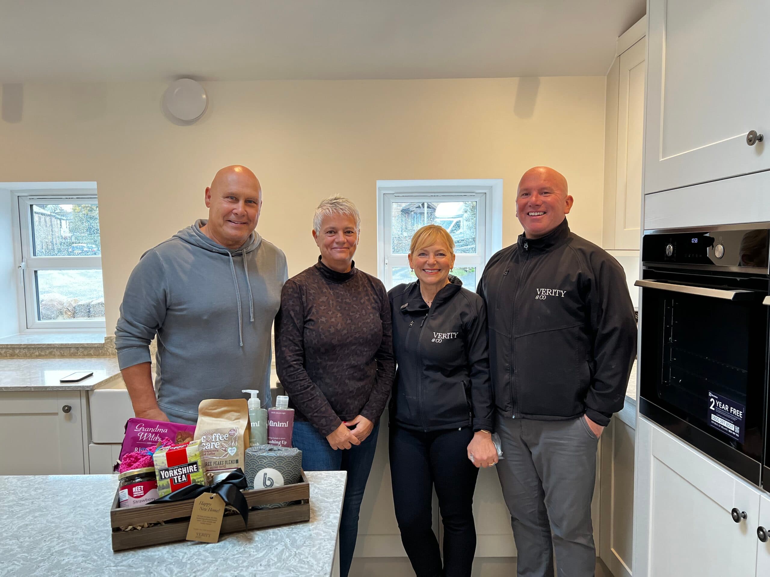 Four people smiling together in the kitchen of a new home at Plot 32, The Laithe, Pendle Farm in Worsthorne, celebrating the handover with a Verity & Co Homes welcome hamper on the kitchen island.